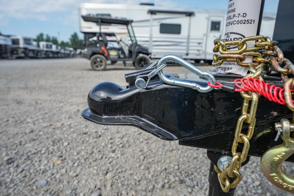 Close-up of a trailer hitch with a metal coupler, safety chains, and a padlock on a gravel lot.