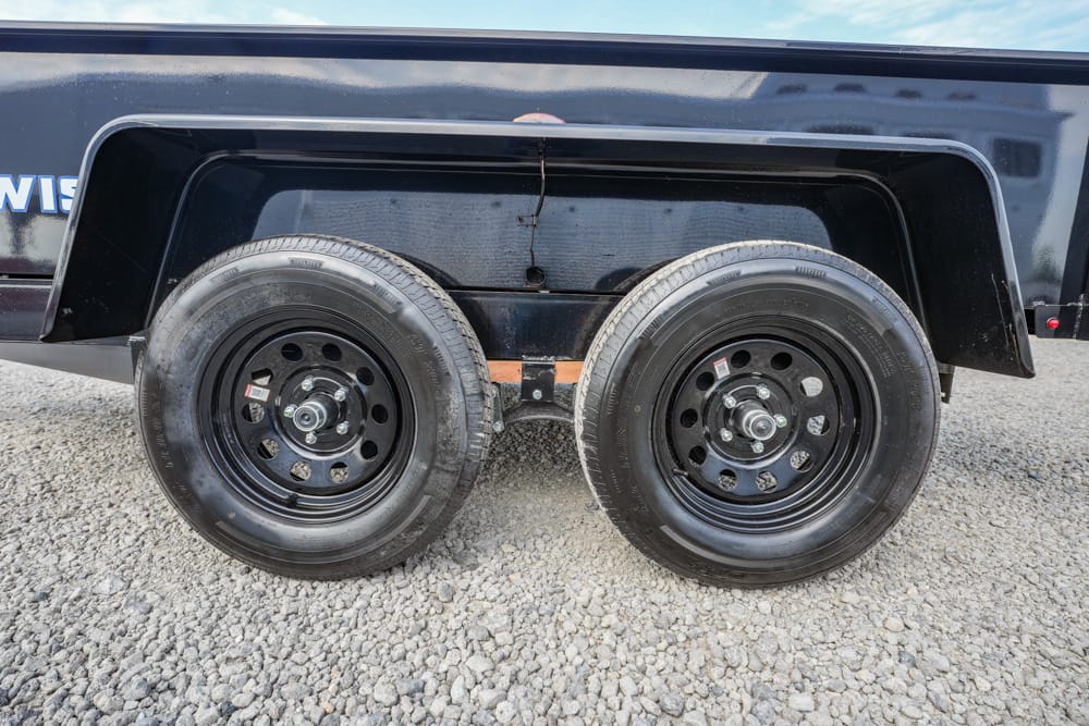 Dual-axle trailer with two black steel wheels on a gravel lot on a sunny day.