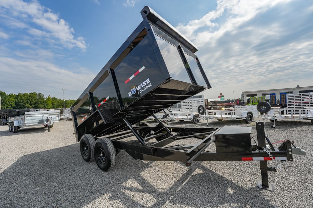 Black tilt-bed dump trailer on a gravel lot, raised to dump position with dual axles visible.