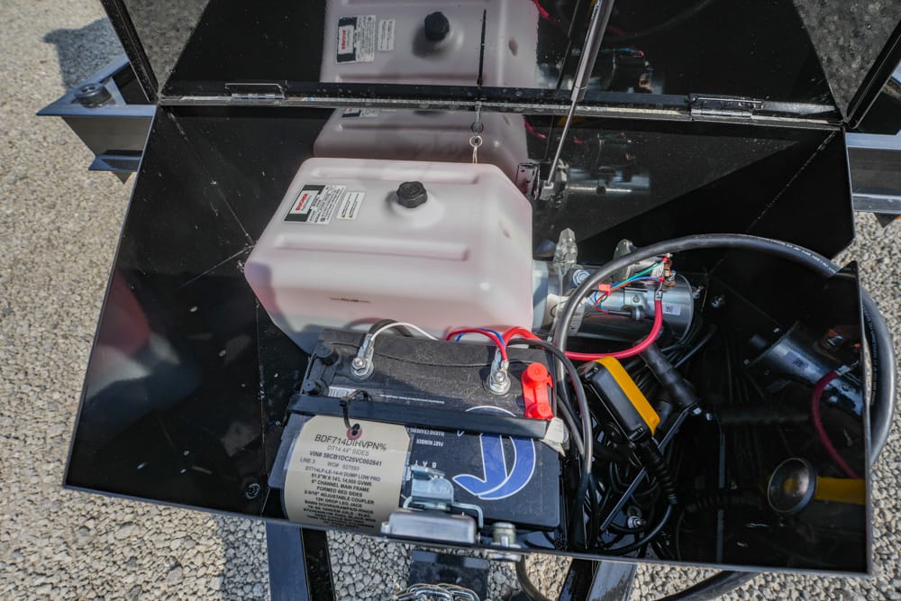 Open equipment compartment showing a car battery, a white plastic reservoir, and tangled wires and hoses inside a black metal case.