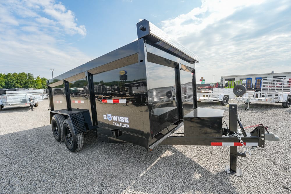 Black dual-axle utility trailer on a gravel lot, with a front hitch and toolbox, labeled B-WISE 7 GA FLOOR in white lettering