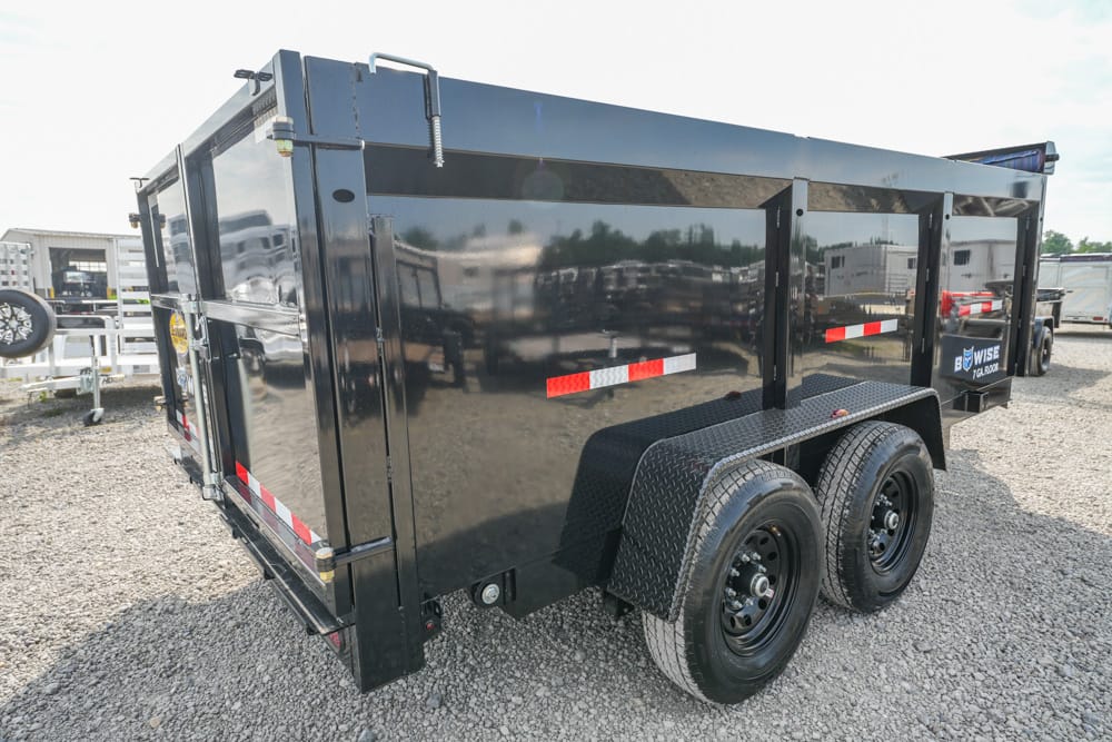 Black dual-axle dump trailer on a gravel lot, showing rear and side panels with reflective stripes.