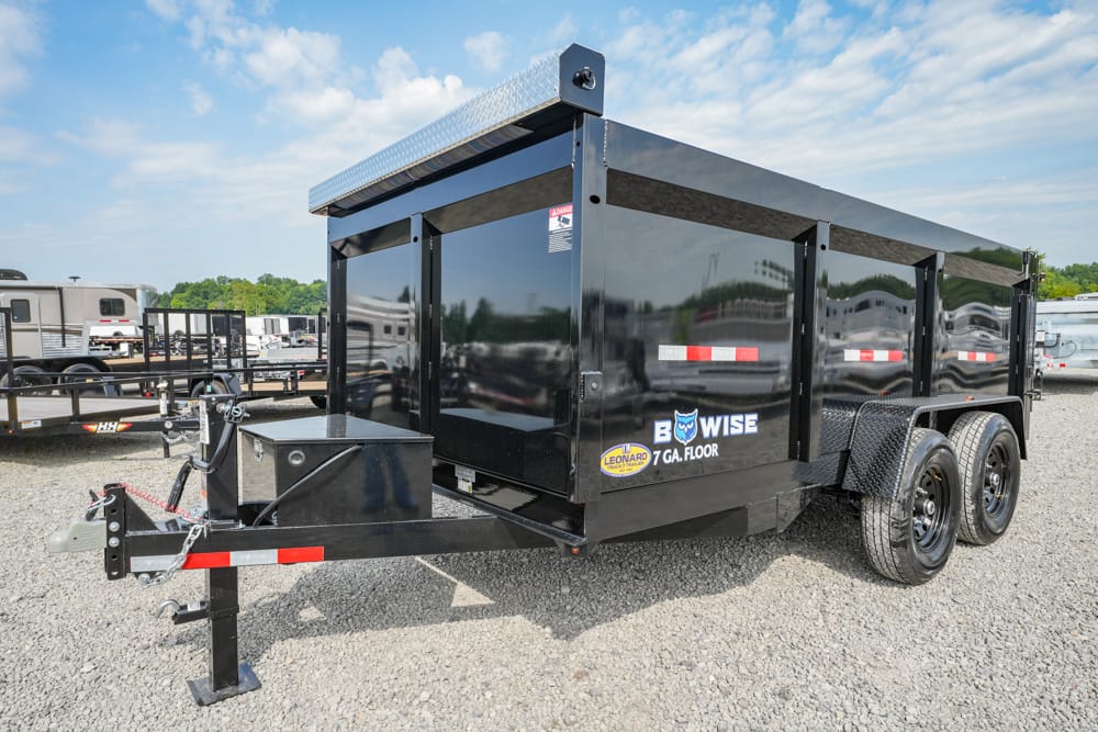 Black tandem-axle dump trailer on a gravel lot, with a hitch, toolbox, and safety chains visible