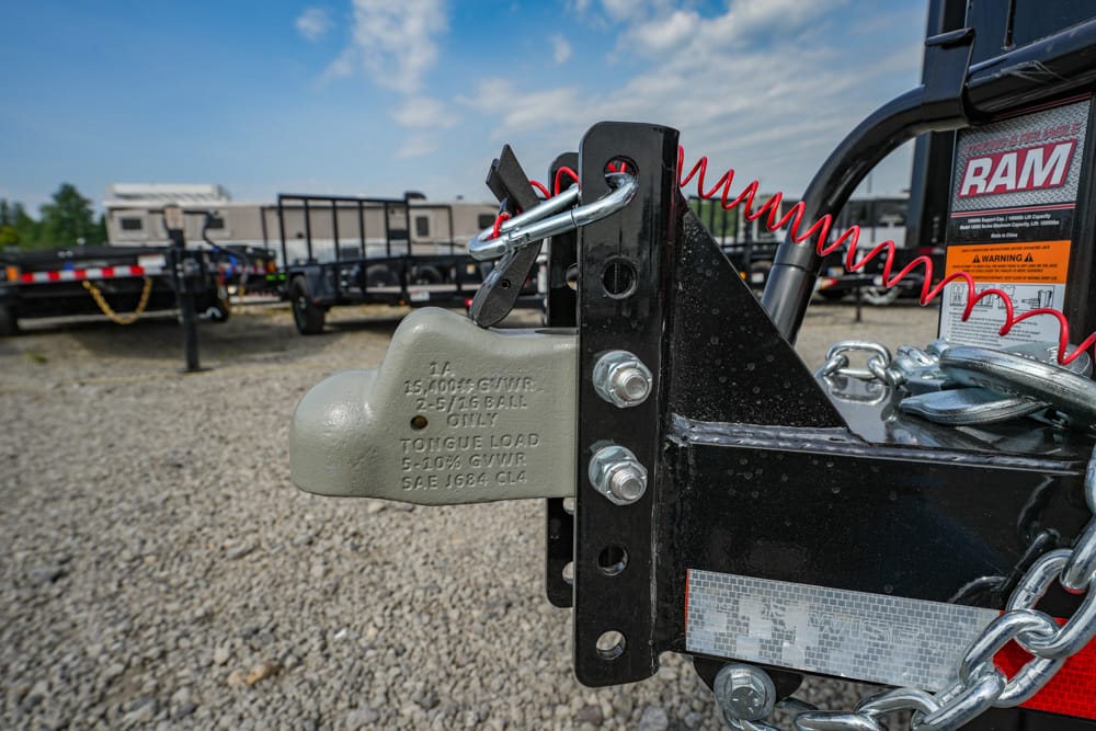 Close-up of a trailer hitch with a grey tongue weight plate, safety chain, and red coiled cord; gravel lot with trailers in the background.