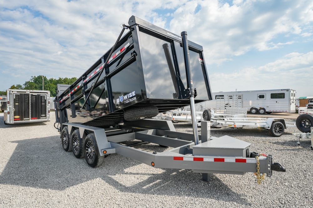 Tilted black dump trailer loaded on a multi-axle gooseneck trailer in a gravel lot, with other trailers and horse trailers in the background.