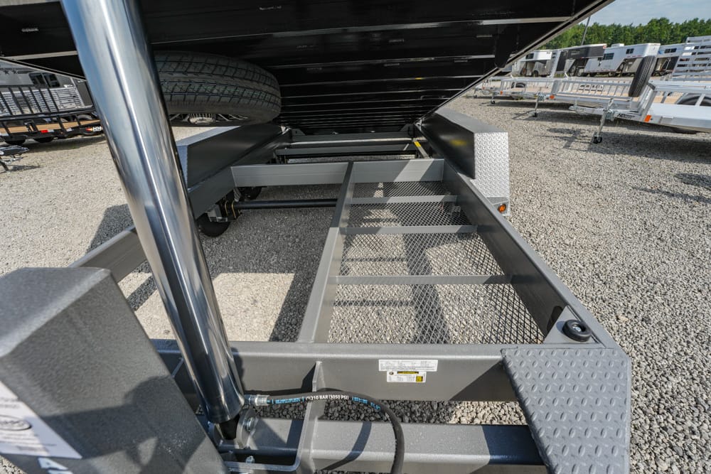 Underneath a flatbed trailer showing a metal mesh ramp/deck, spare tire, and gravel lot with other trailers in the background.