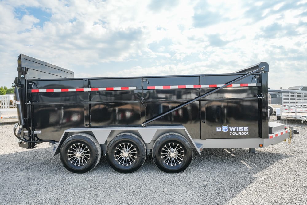 Side view of a black three-axle dump trailer with a glossy finish, red reflective stripes, and brand 'B-WISE 7 GA. FLOOR' on a gravel lot under a blue sky with clouds