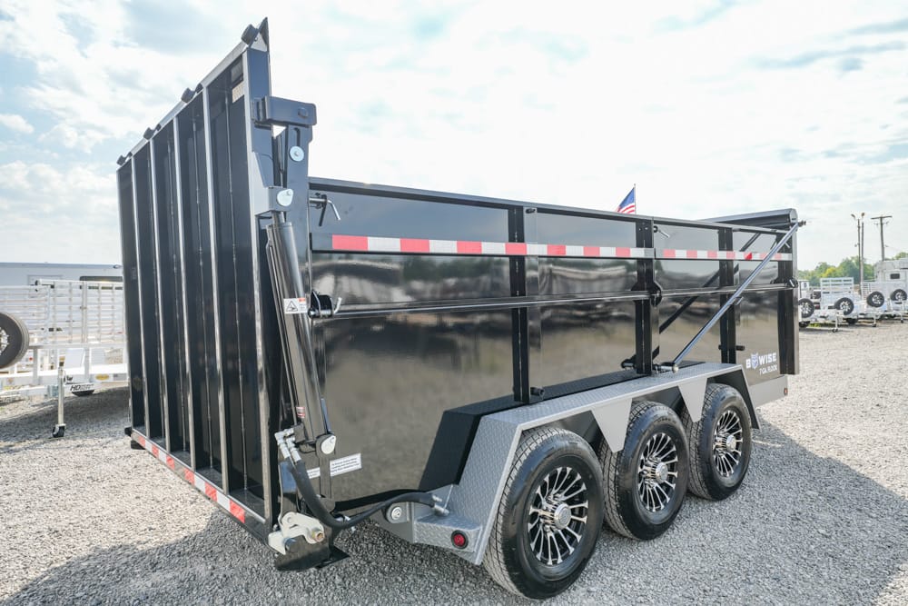 Black tri-axle dump trailer with diamond-plate fenders parked on a gravel lot, side view.
