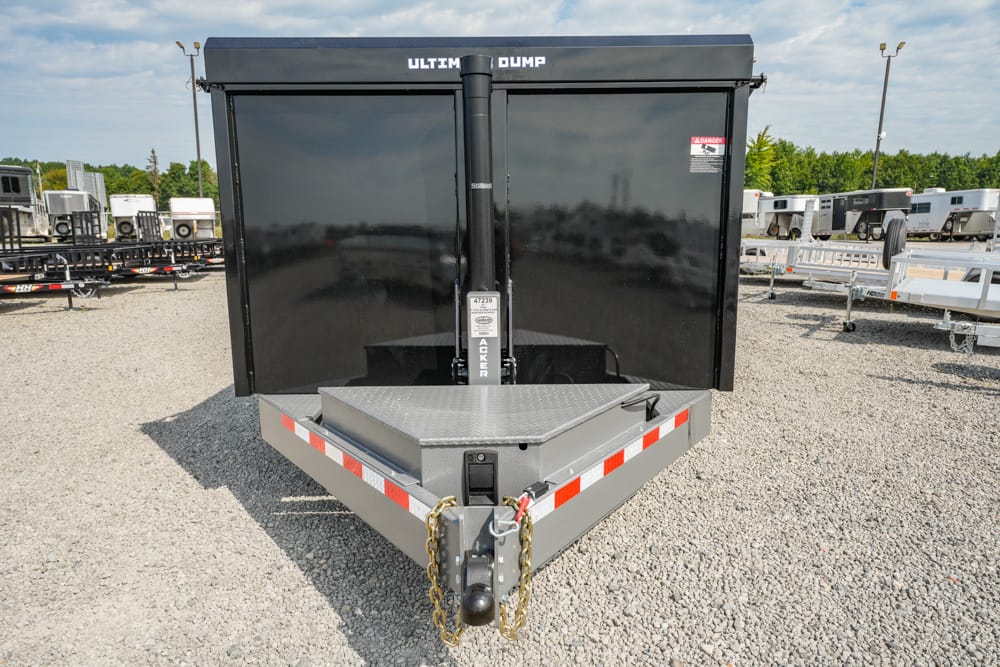 Front view of a black Ultimate Dump trailer with a diamond-plate step and safety chains, parked on a gravel lot.