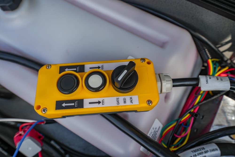 Yellow control pendant with three round buttons and a toggle switch, mounted on a surface with multi‑colored wires in the background; labels suggest controls for gate, bed, and jack.