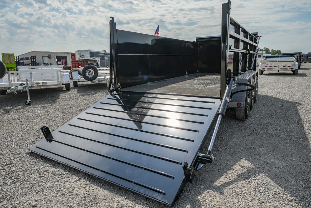 Black dump trailer with lowered metal ramp on a gravel lot, other trailers in the background and an American flag in the sky.