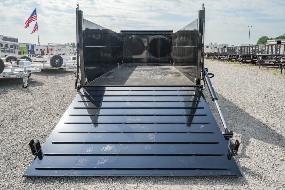 Black dump trailer with its open bed and ramp on a gravel trailer yard, with other trailers and an American flag in the background.