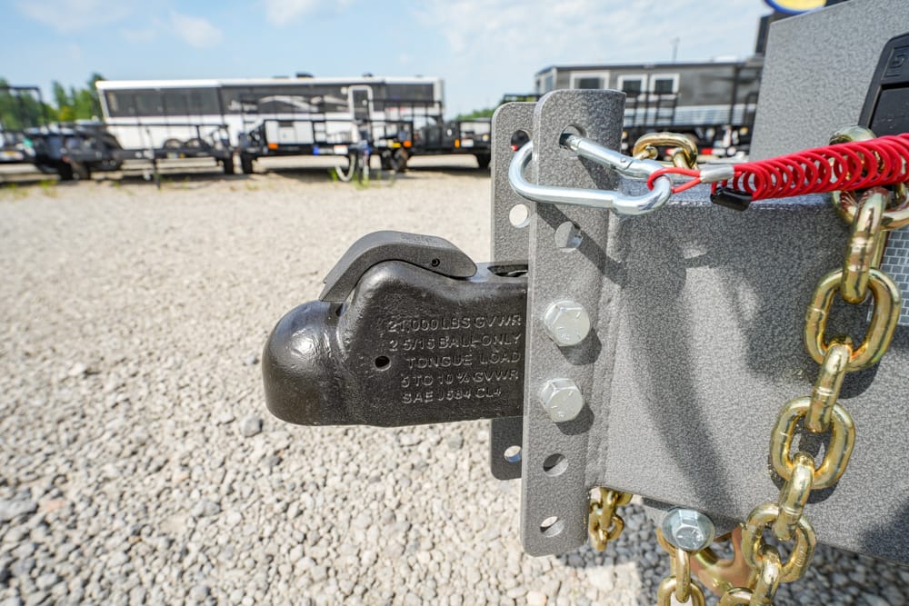Close-up of a trailer hitch with a black coupler attached to a gray steel frame, safety chain loops, and a red coiled safety wire against a gravel lot background.