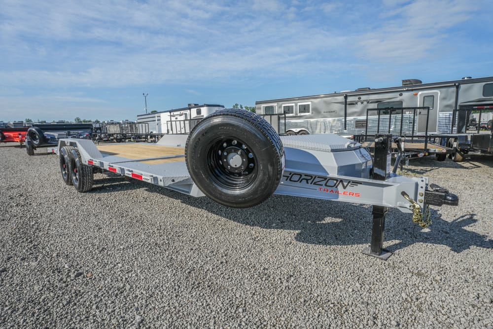 Long, flatbed Horizon trailer with a spare tire mounted on the front, parked on a gravel lot with other trailers and RVs in the background.