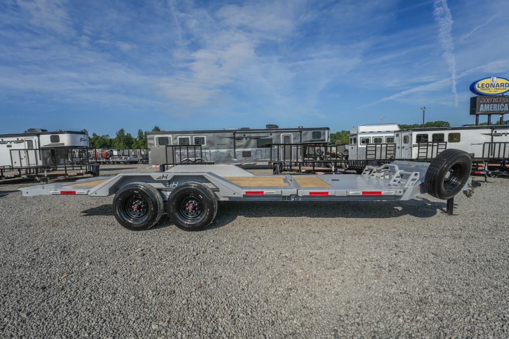 Silver car hauler trailer with dual rear axles and a mounted spare tire at the front, parked on gravel with RVs and trailers in the background.