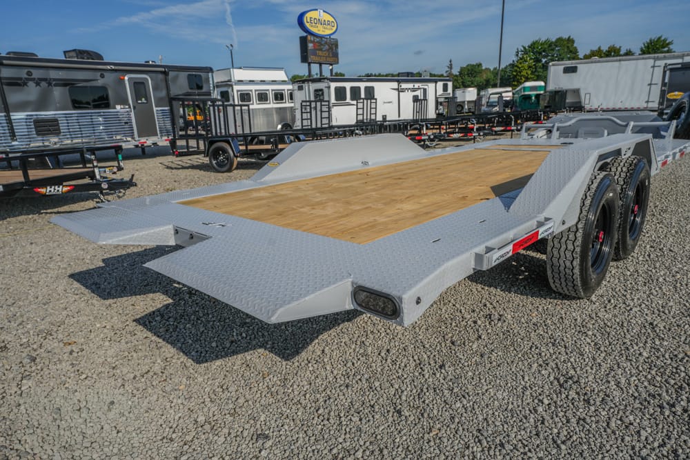Silver steel flatbed trailer with a wooden deck and diamond-plate edges, twin axles, on a gravel lot with other trailers nearby.