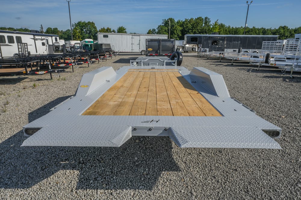 Wide view of a heavy-duty flatbed trailer with a wooden deck and diamond-plate ramps, parked on a gravel lot among other trailers.