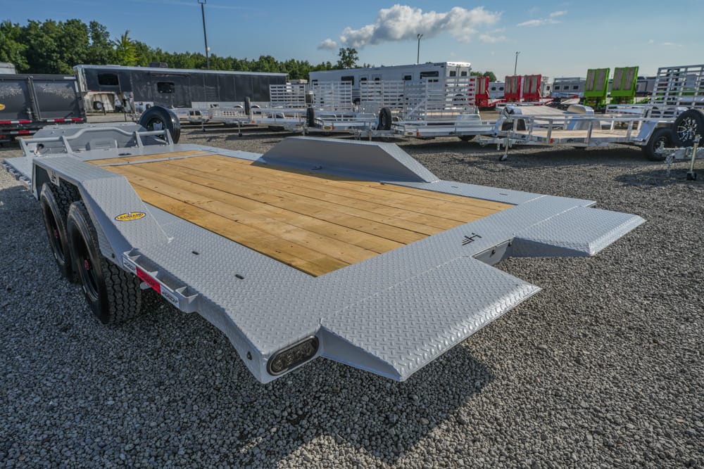 Wide view of a silver diamond-plate flatbed trailer with a wooden deck, dual axles, and ramp extensions, parked on a gravel lot with other trailers in the background