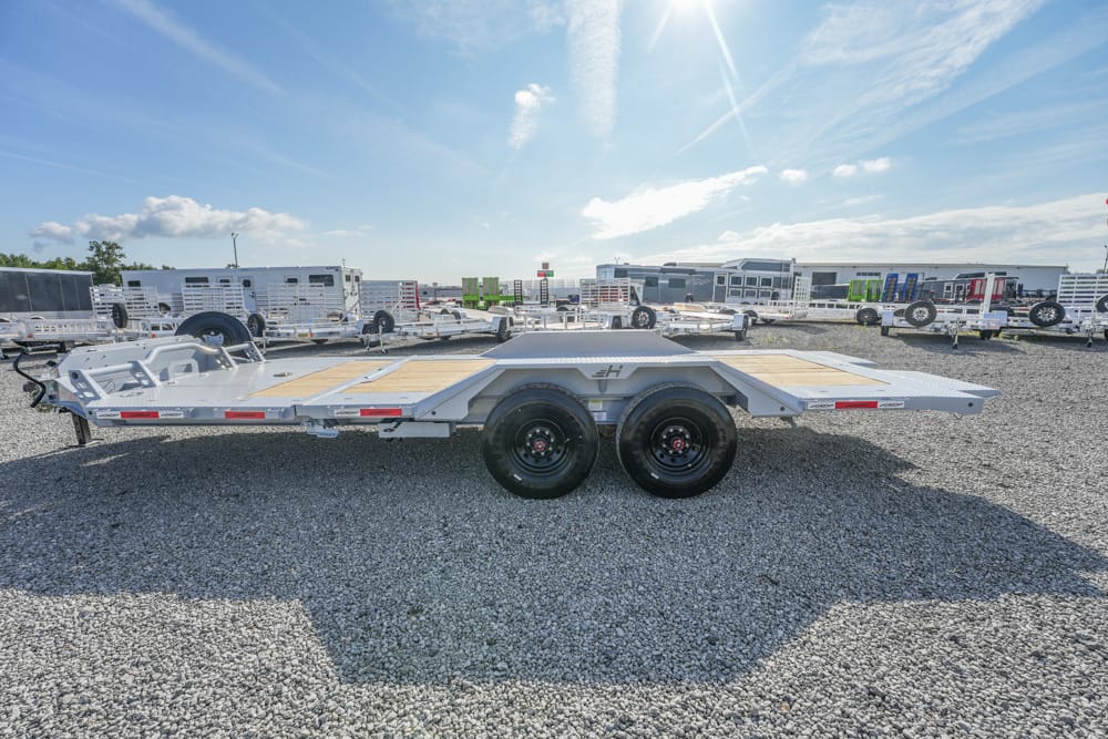 Silver dual-axle car trailer with a wooden deck on a gravel lot, rows of trailers in the background under a blue sky.