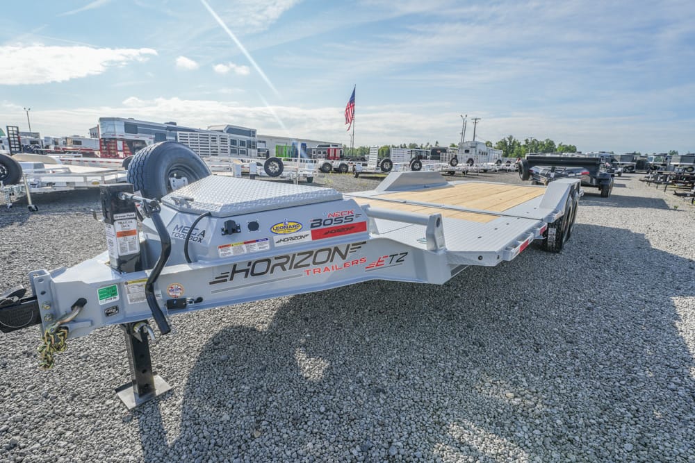 Flatbed utility trailer with wood deck and spare tire, on a gravel lot at a trailer dealership under a blue sky with other trailers nearby.