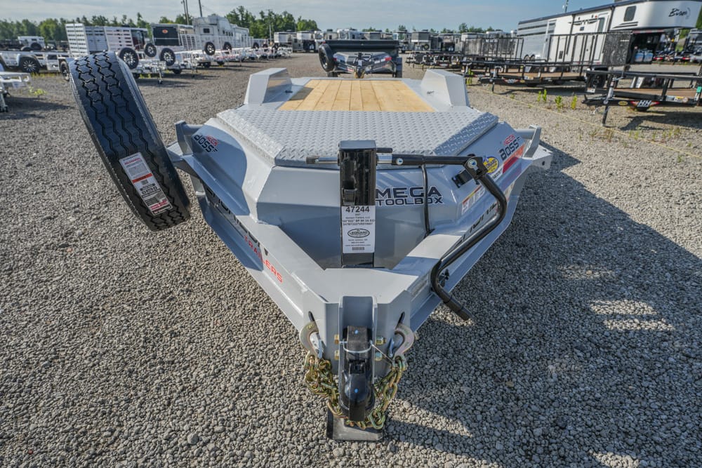 Front view of a gray heavy-duty equipment trailer on a gravel lot, spare tire mounted on the left and a wooden deck visible at the rear.