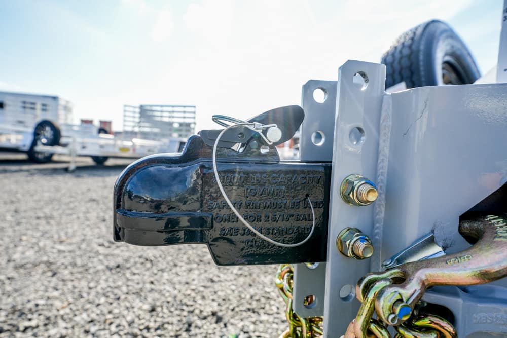 Close-up of a black trailer hitch coupler secured to a gray trailer frame with a safety pin and chain links visible, ready for towing.