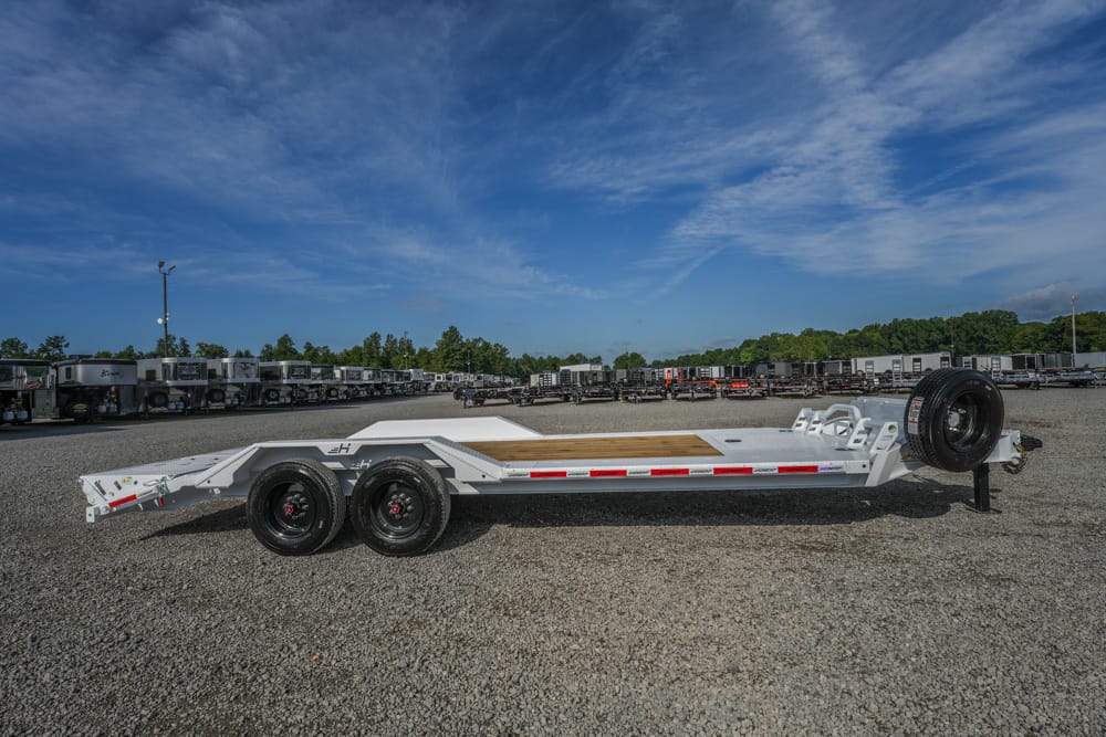 White flatbed car-hauler trailer with dual rear axles and a spare tire, parked on a gravel lot with numerous trailers in the background under a blue sky.