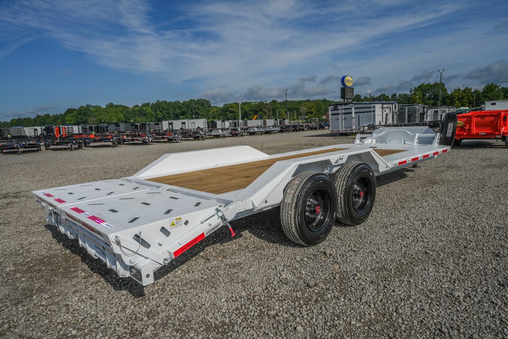 White heavy-duty tilt-bed trailer with two-axle rear set, wooden deck, parked on a gravel lot with numerous trailers in the background.