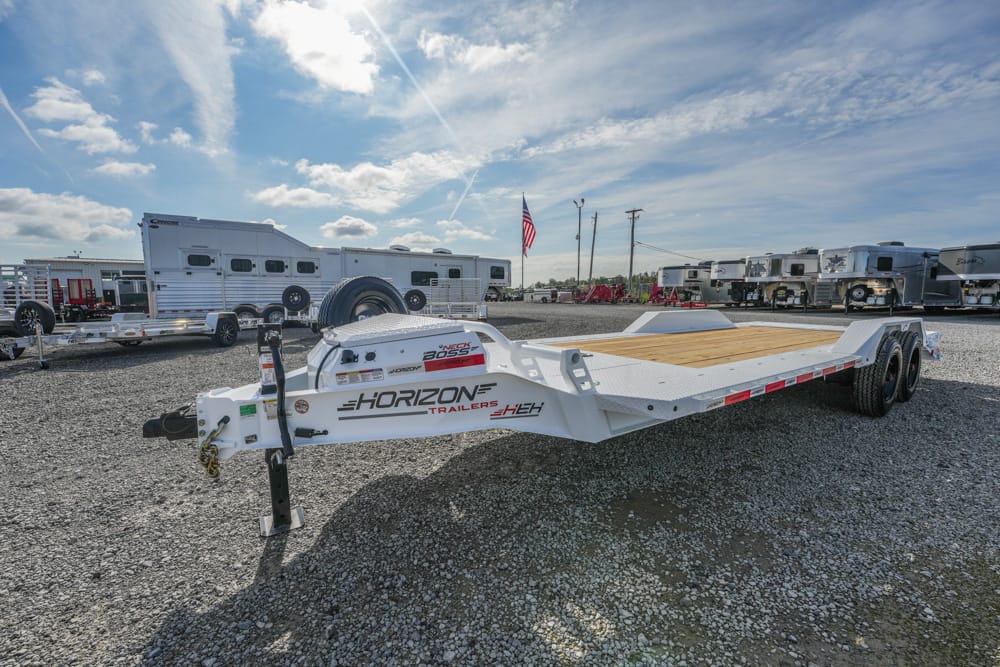White Horizon car trailer with wooden deck in a gravel lot, surrounded by RVs and trailers; American flag in background.