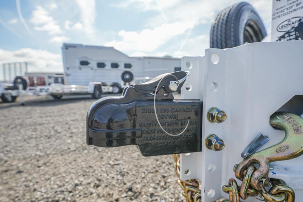 Close-up of a black trailer coupler with a latch on a white trailer frame, safety chains looping nearby, and trucks in the background.