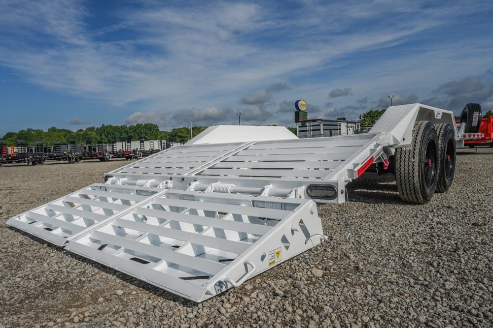 White heavy-duty trailer with ramp sections lowered on a gravel lot, other trailers visible in the background under a blue sky.