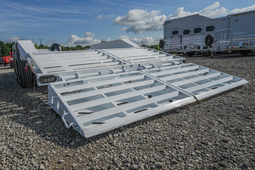 White metal car-hauler trailer with folded loading ramps on a gravel lot and other trailers in the background under a blue sky.