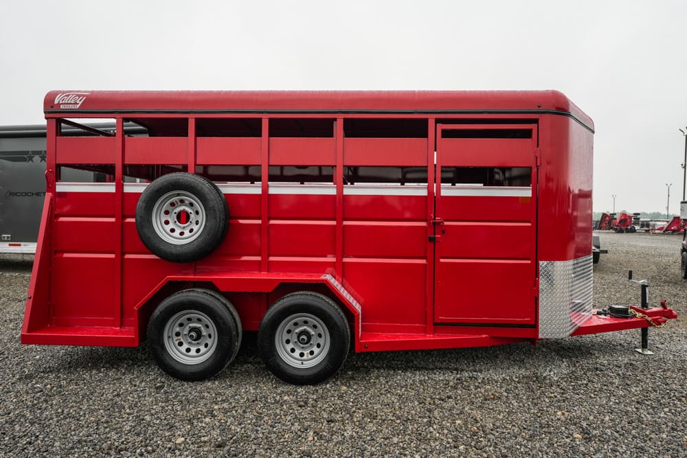 Red double-axle livestock/horse trailer with a mounted spare tire on its side, parked on gravel