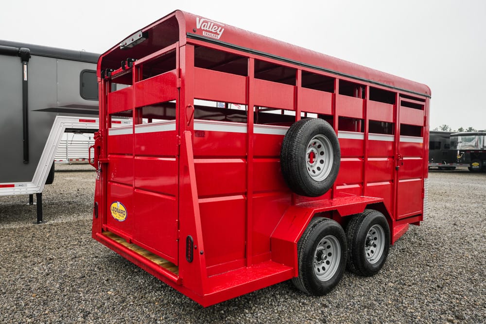 Red dual-axle livestock trailer with a side-mounted spare tire, parked on a gravel lot