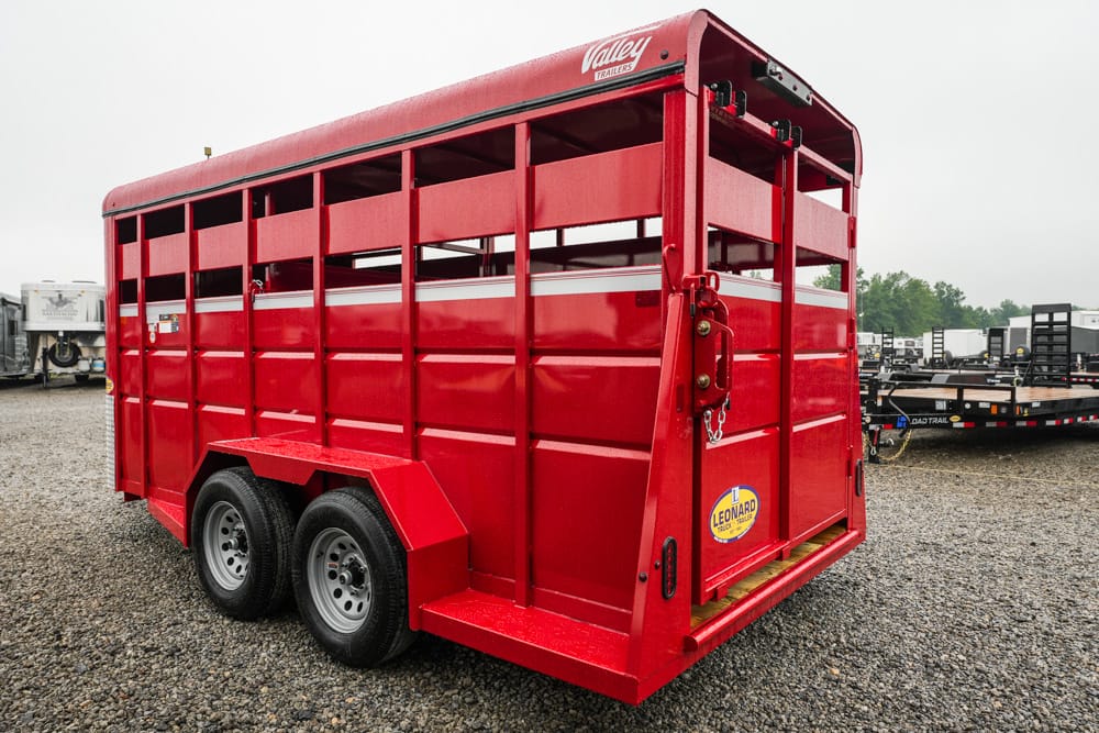 Red dual-axle livestock trailer on a gravel lot, with a Leonard sticker on the back and open slat sides partially visible behind the hitch.