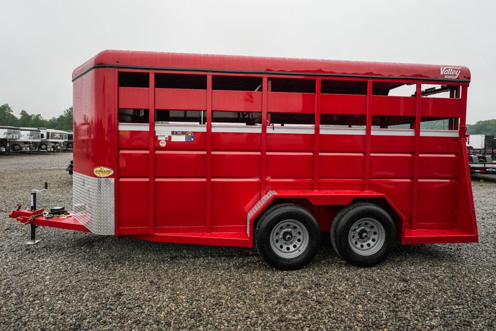 Red livestock trailer with tandem wheels parked on a gravel lot, ready for hauling animals on a hitch.