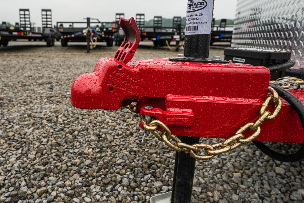 Red trailer hitch with a gold chain looped around it, covered in raindrops, on a gravel lot with trailers in the background.