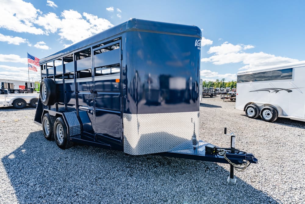 Blue gooseneck horse trailer with diamond-plate front, parked on gravel lot with other trailers and an American flag in the background.