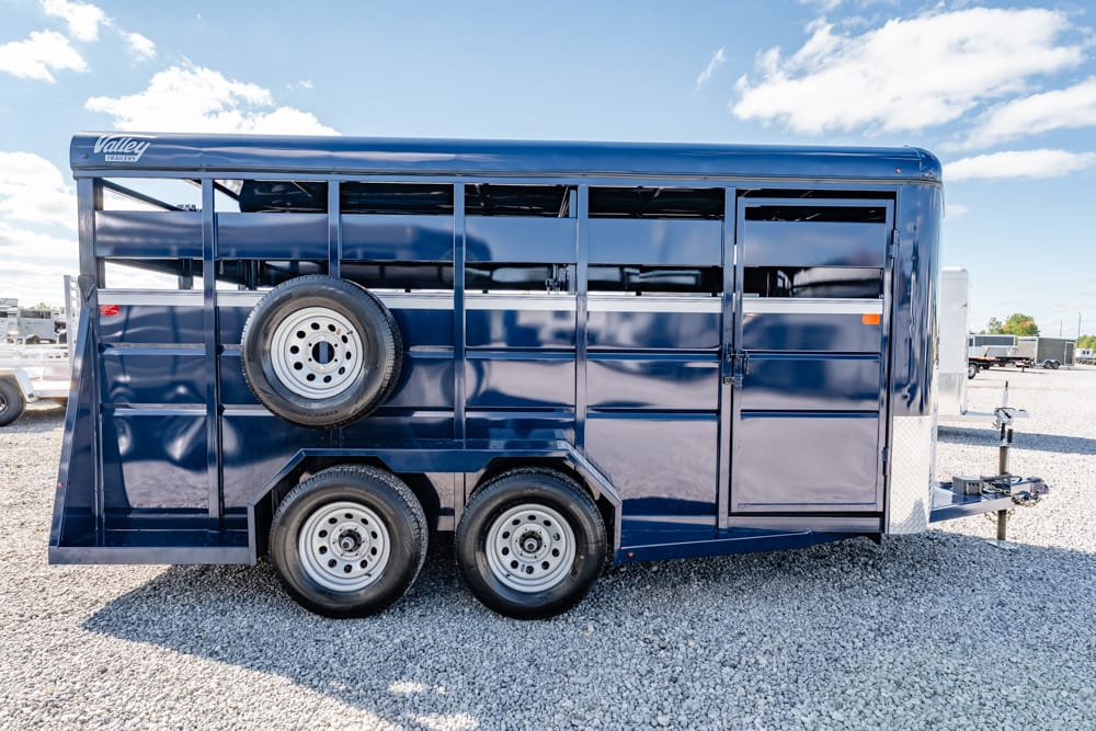 Blue livestock trailer with two axles and a spare tire mounted on the side, parked on a gravel lot under a sunny sky