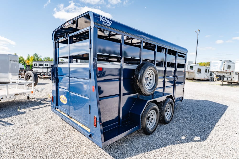Blue livestock trailer with three axles and a side-mounted spare tire on a gravel lot.