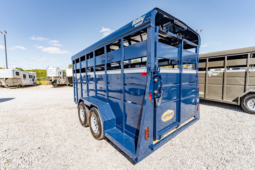 Rear view of a blue double-axle livestock trailer on a gravel lot, with other trailers visible in the background.