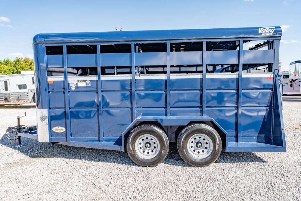 Blue metal livestock trailer with two axles and vented side rails parked on a gravel lot on a sunny day.