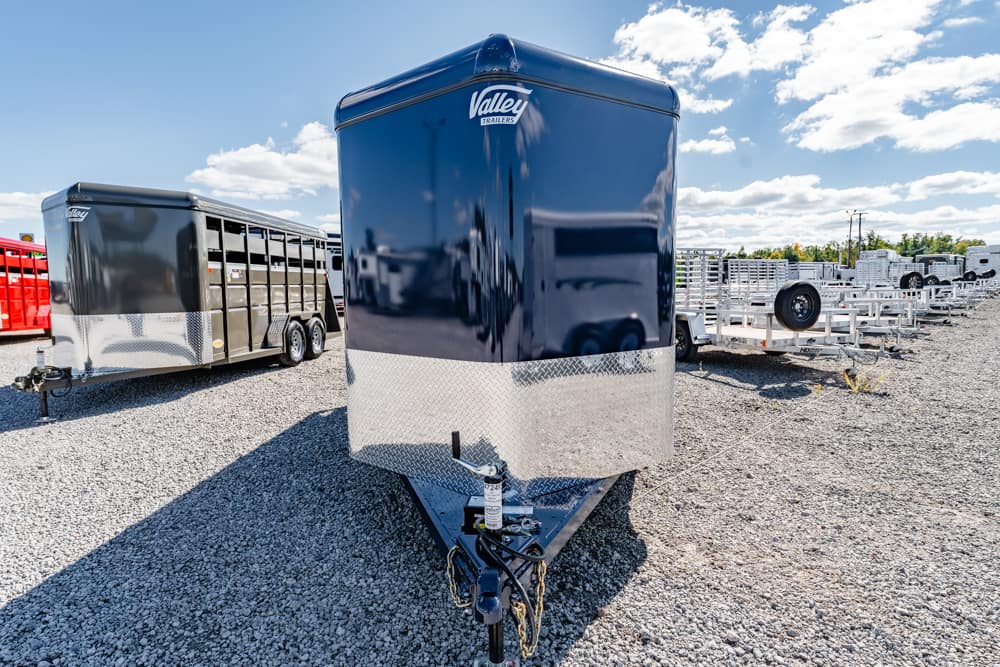 Blue enclosed trailer with a diamond-plate front sits on a gravel lot, other trailers visible in the background, under a sunny sky.