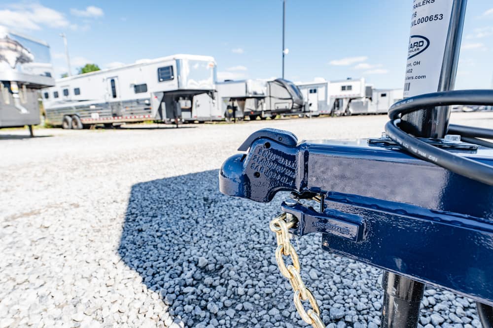 Close-up of a blue trailer hitch with a gold chain, parked RVs in a gravel lot under a bright blue sky.