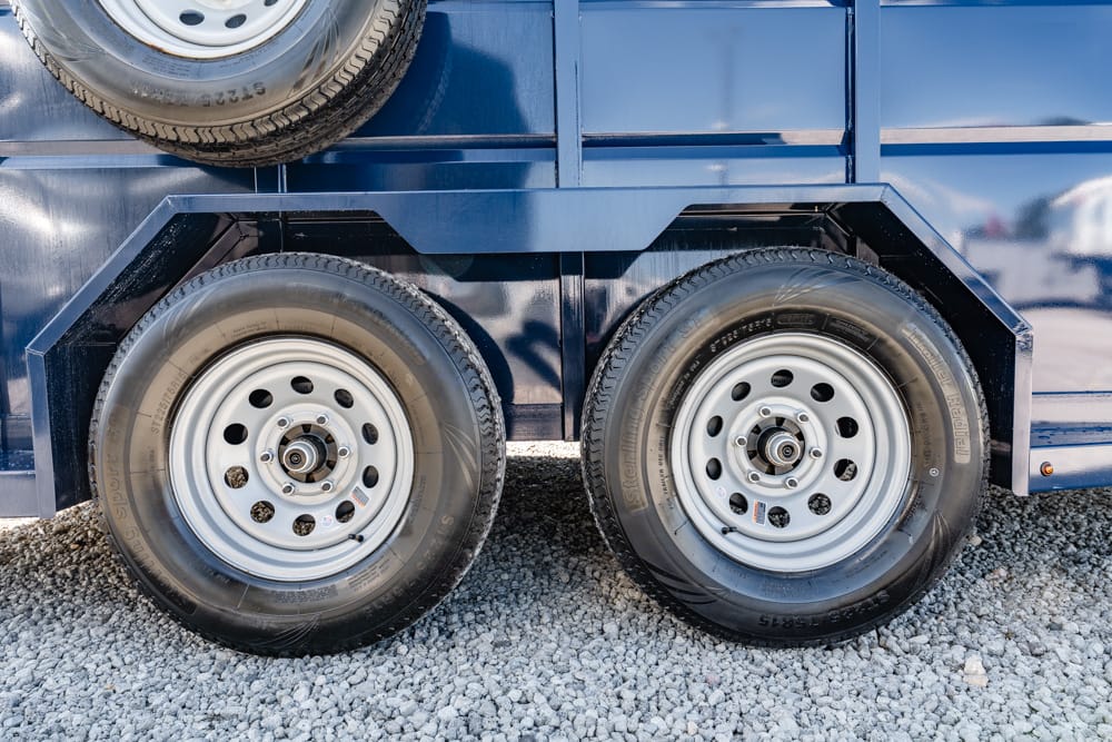 Blue utility trailer with two rear axles and dual tires on gravel, showing wheel rims and spare tire above.