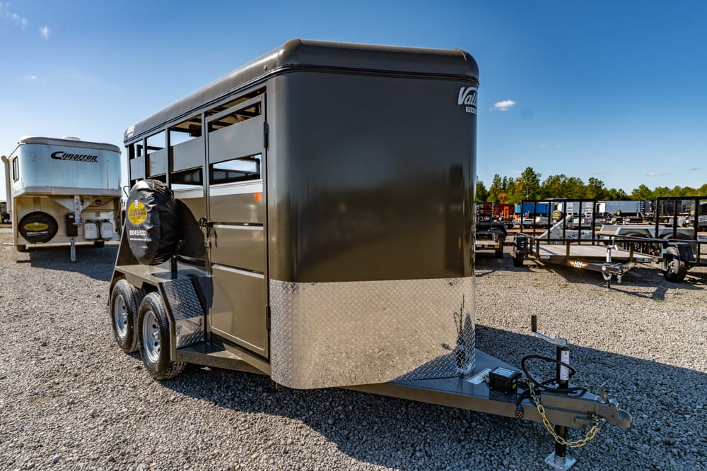 Black, enclosed trailer with dual axles and diamond-plate front in a gravel lot, ready to tow. The spare tire is mounted on the side.