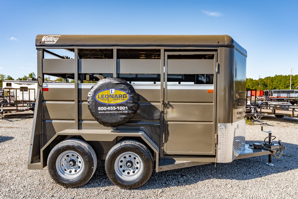 Brown dual-axle horse trailer with a Leonard Truck Trailer spare tire cover, parked on gravel with a clear blue sky