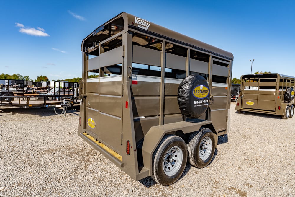 Brown metal livestock trailer with a spare tire cover on the back, parked on a gravel lot among other trailers.