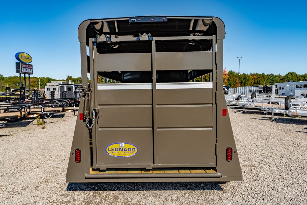 Rear view of a brown livestock trailer with its back doors closed, Leonard logo on the rear, parked on a gravel lot with other trailers nearby.