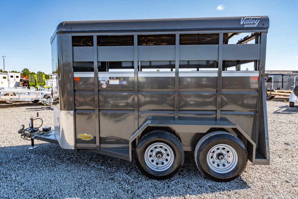 Gray double-axle livestock trailer parked on a gravel lot, front hitch visible, Valley Trailers logo on top corner, side panels with vents and a diamond plate at the front lower edge
